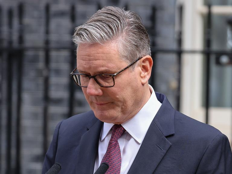 Keir Starmer steht im Anzug mit gesenktem Blick vor einem Metallzaun