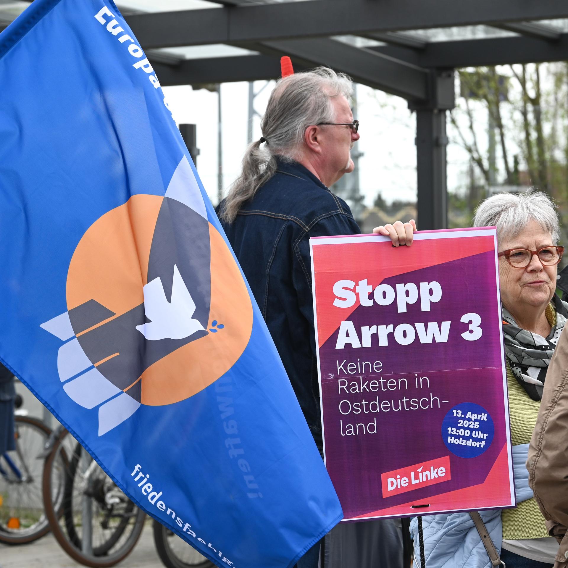Demonstranten protestieren mit Fahnen und Transparenten gegen die Raketenstationierung bei Holzdorf. Auf einem Plakat steht "Stopp Arrow 3 – keine Raketenstationierung in Ostdeutschland!"