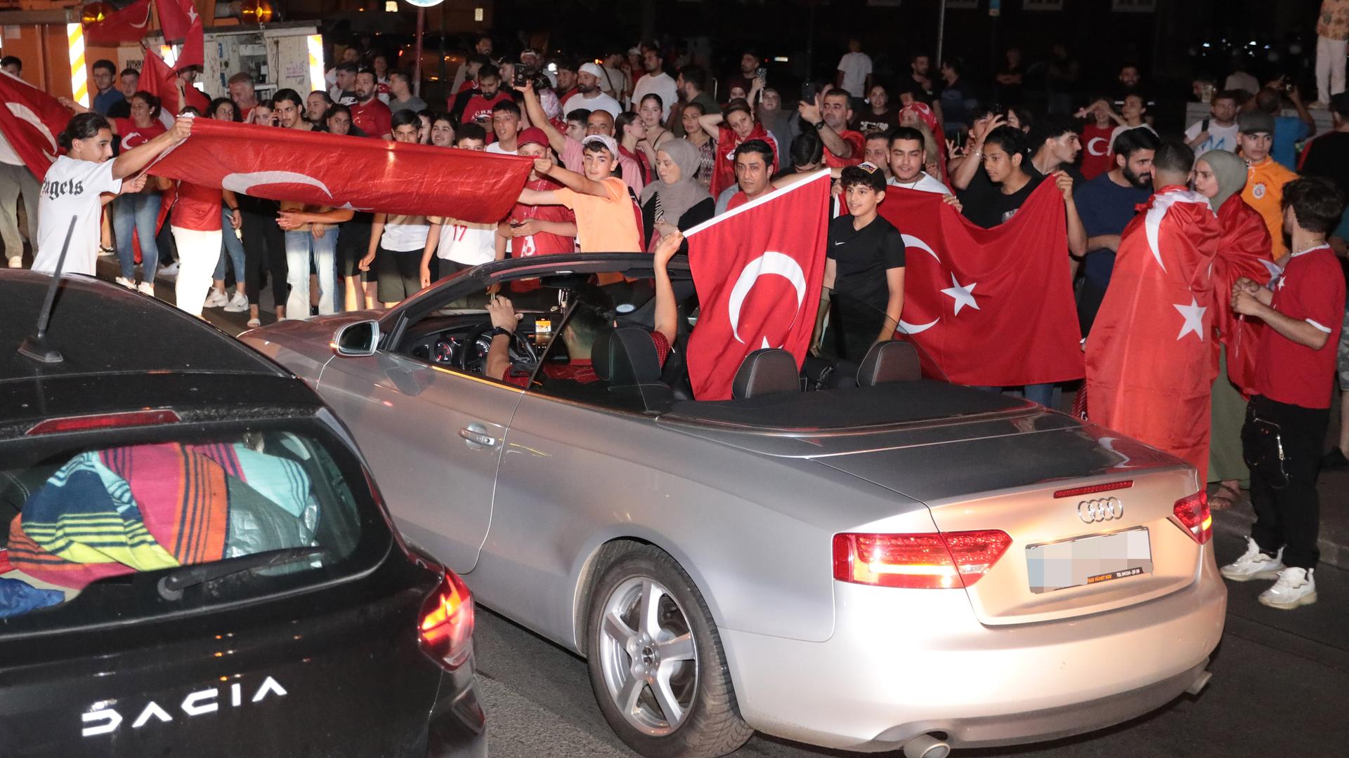 Jubelnde Fans mit türkischen Flaggen umringen ein silberfarbenes Audi-Cabriolet und einen schwarzen Dacia-Kleinwagen Jubelnde Fans mit türkischen Flaggen umringen ein silberfarbenes Audi-Cabriolet und einen schwarzen Dacia-Kleinwagen