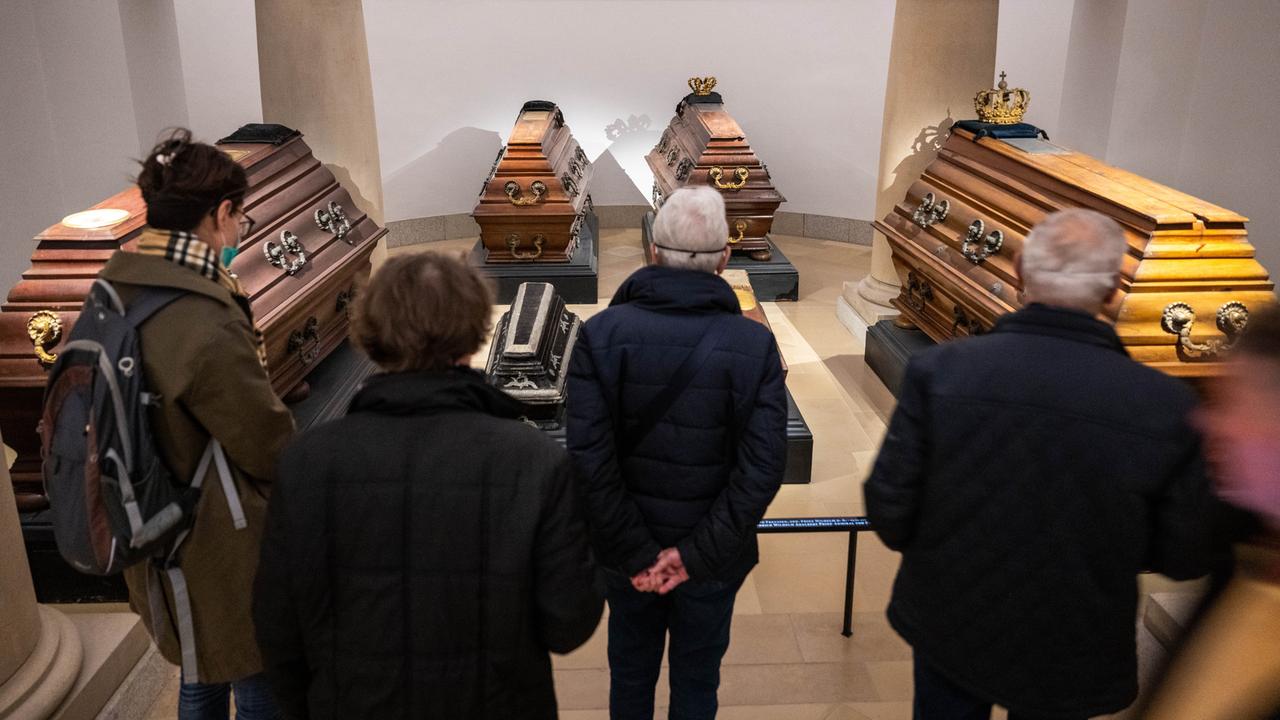 Mehrere Menschen stehen vor einer Absperrung, dahinter stehen vier hölzerne Särge mit Verzierungen.