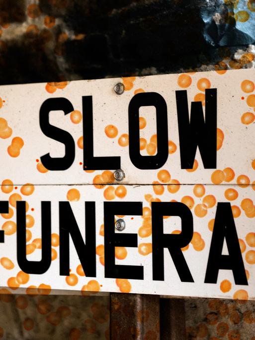 Ein Schild mit der Aufschrift "Slow Funeral". Die Fotografie wird durch eine Grafik von orange farbenen Punkten überlagert.