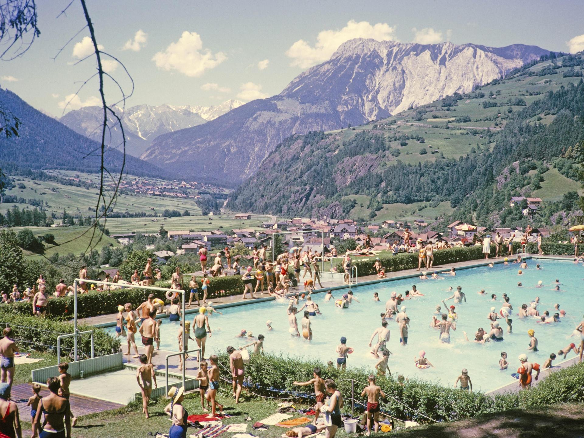 Gut gefülltes Schwimmbad vor der Kulisse der Alpen an einem sonnigen Sommertag Ötz in Österreich. Historische, kolorierte Postkarte.