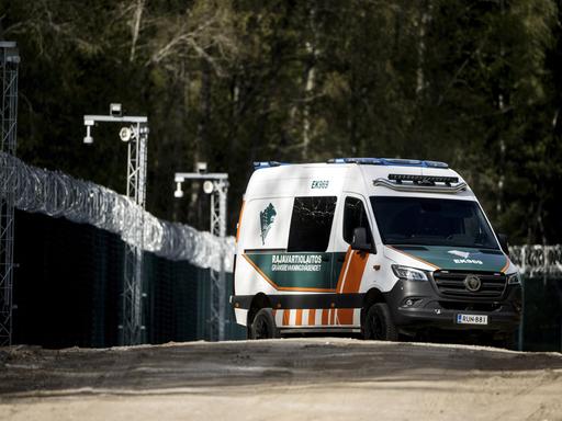 Ein finnisches Grenzschutzfahrzeug fährt auf einer unbefestigten Straße entlang eines hohen Zauns mit Stacheldraht. Im Hintergrund stehen Bäume und Überwachungskameras.