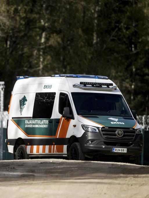 Ein finnisches Grenzschutzfahrzeug fährt auf einer unbefestigten Straße entlang eines hohen Zauns mit Stacheldraht. Im Hintergrund stehen Bäume und Überwachungskameras.