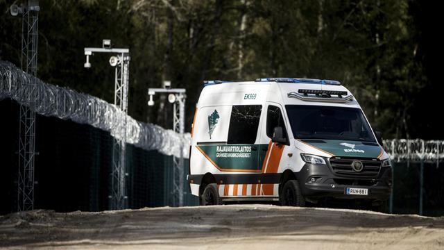 Ein finnisches Grenzschutzfahrzeug fährt auf einer unbefestigten Straße entlang eines hohen Zauns mit Stacheldraht. Im Hintergrund stehen Bäume und Überwachungskameras.