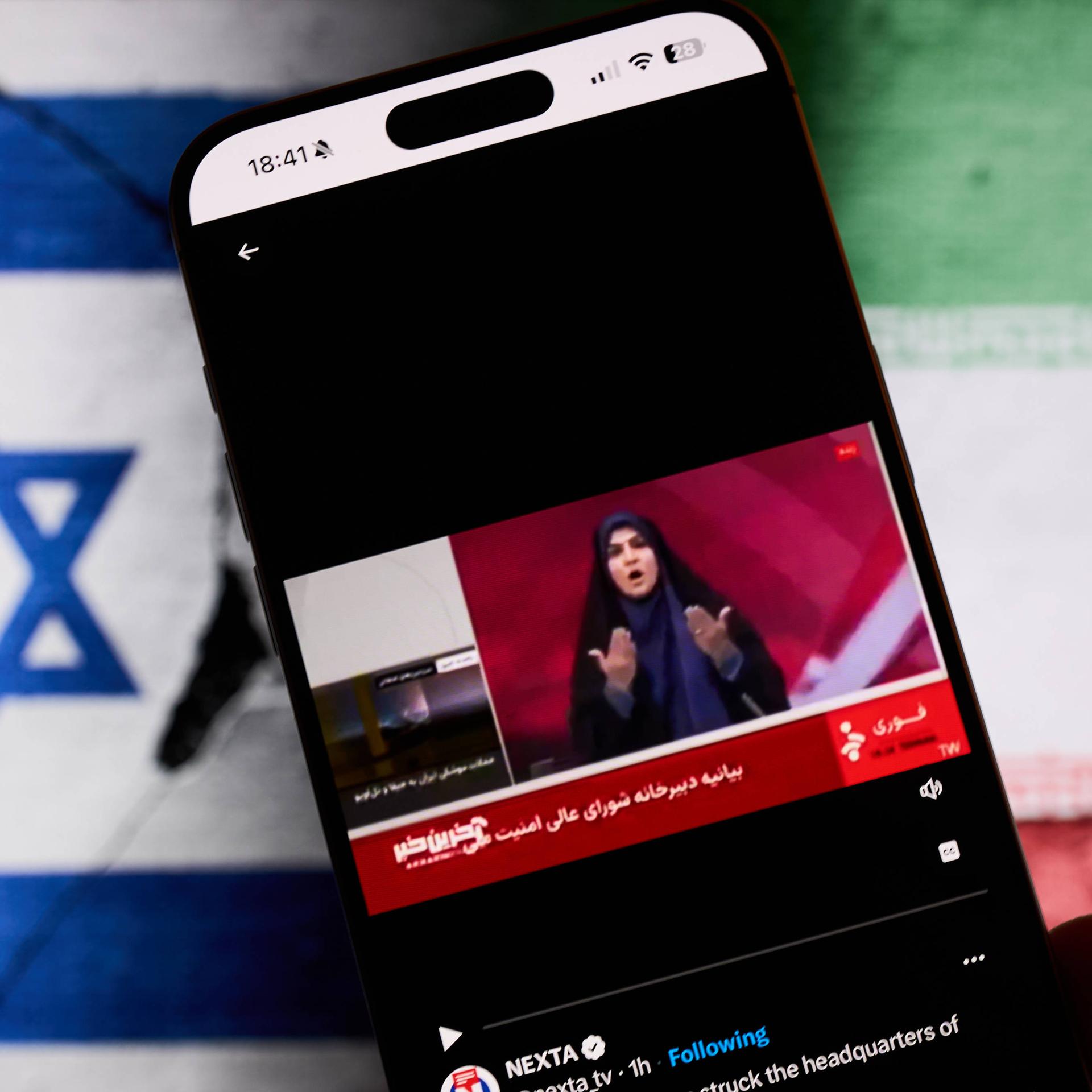 Fotomontage: Im Hintergrund die iranische und israelische Flaggen. Im Vordergrund ein Mobiltelefon: darauf abgeildet ist die Moderatorin des staatlichen Fernsehen im Iran vor einem israelischen Angriff