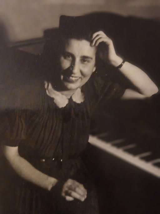Ein schwarz-weiß Foto. Eine junge Frau sitzt vor einem Flügel, stützt den Ellenbogen auf das Instrument und lächelt in die Kamera. Sie trägt kurze, schwarze Haare.