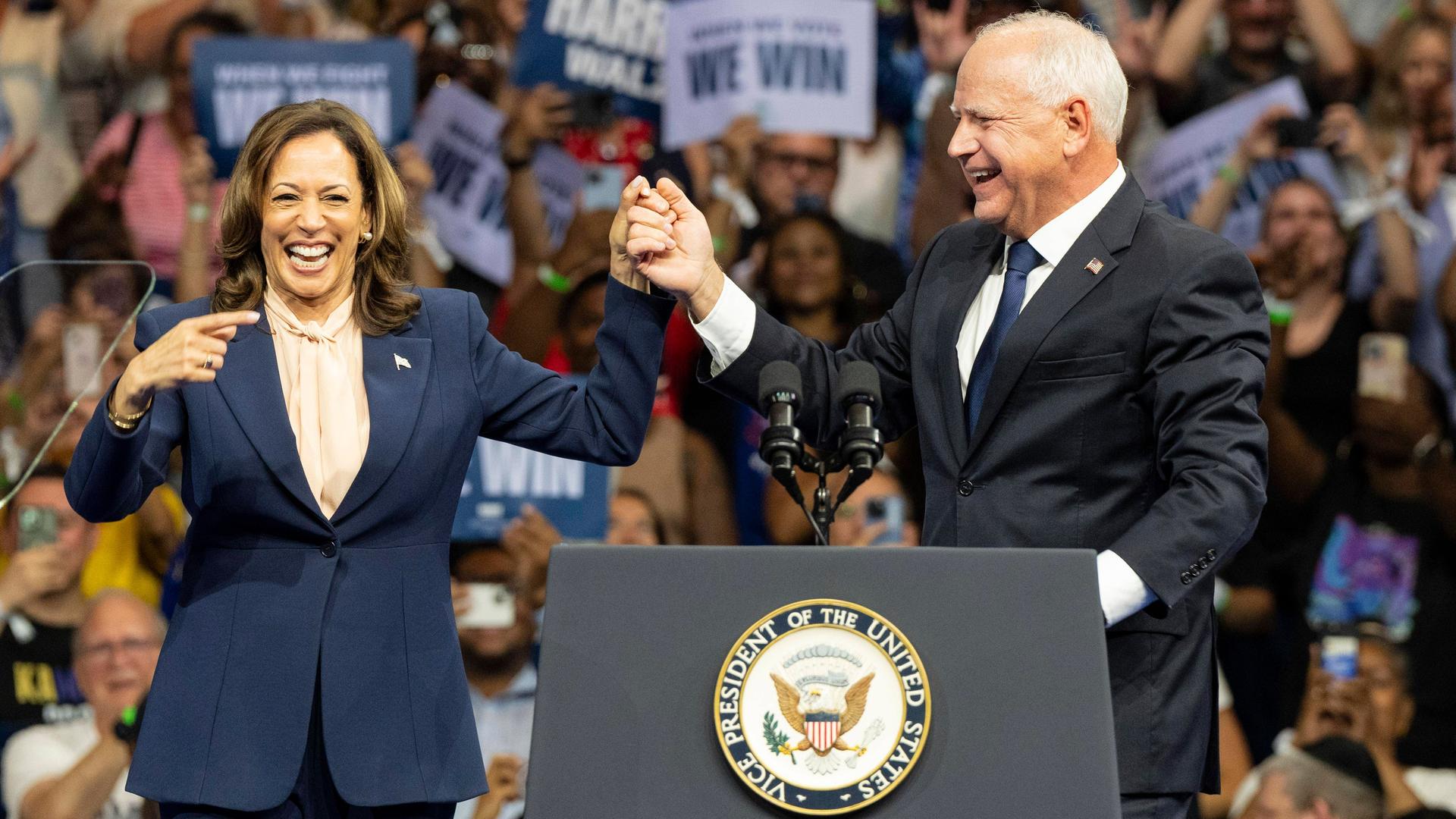 Kamala Harris und Tim Walz stehen während einer Wahlkampf-Veranstaltung auf einer Bühne und lachen.