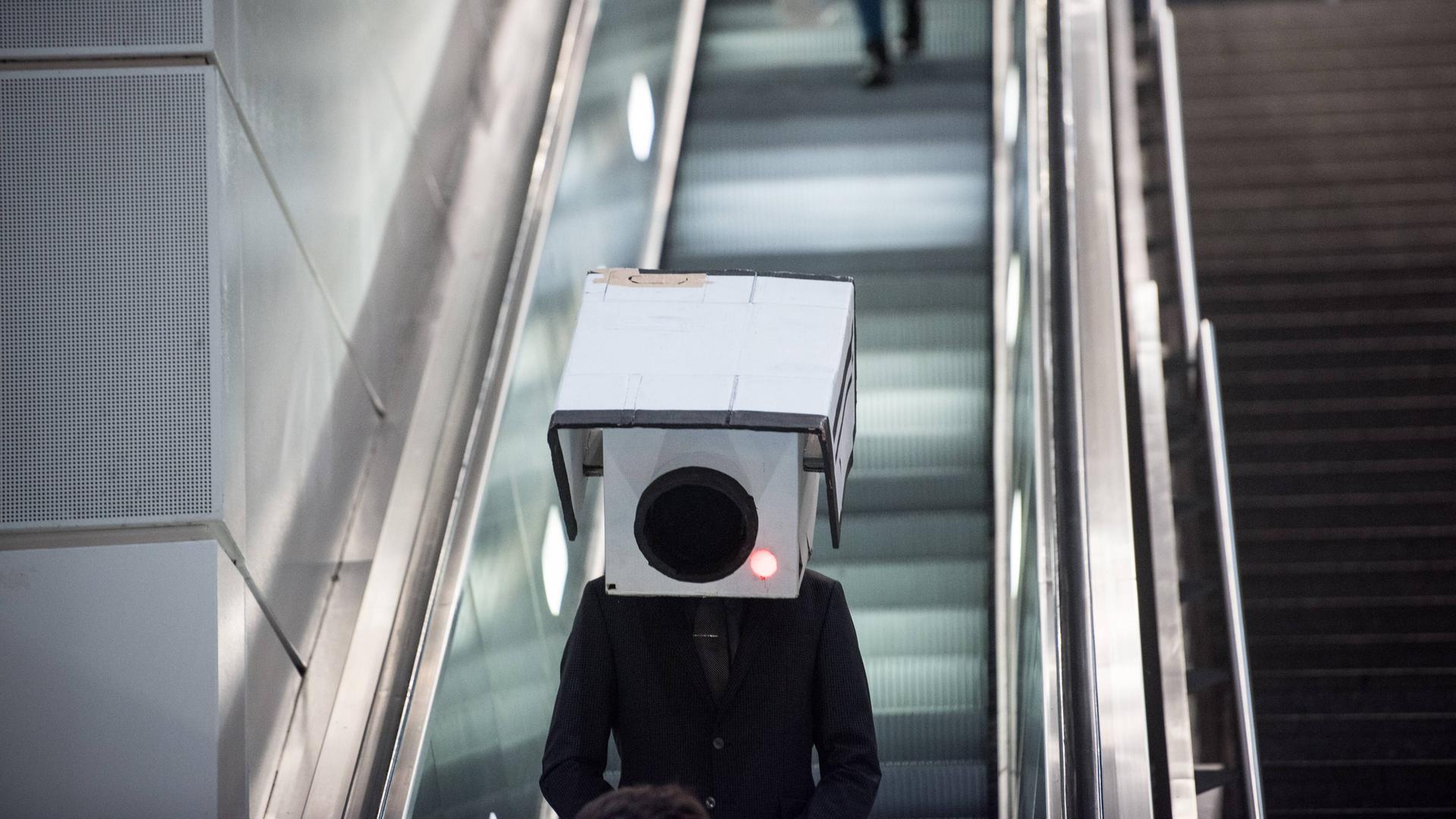 Ein Teilnehmer mit einer Maske in Form einer Überwachungskamera bei einer Aktion gegen den Ausbau von Videoüberwachung im öffentlichen Raum am Bahnhof Südkreuz in Berlin. Die Person trägt einen dunkeln Anzug und steht auf einer Rolltreppe.