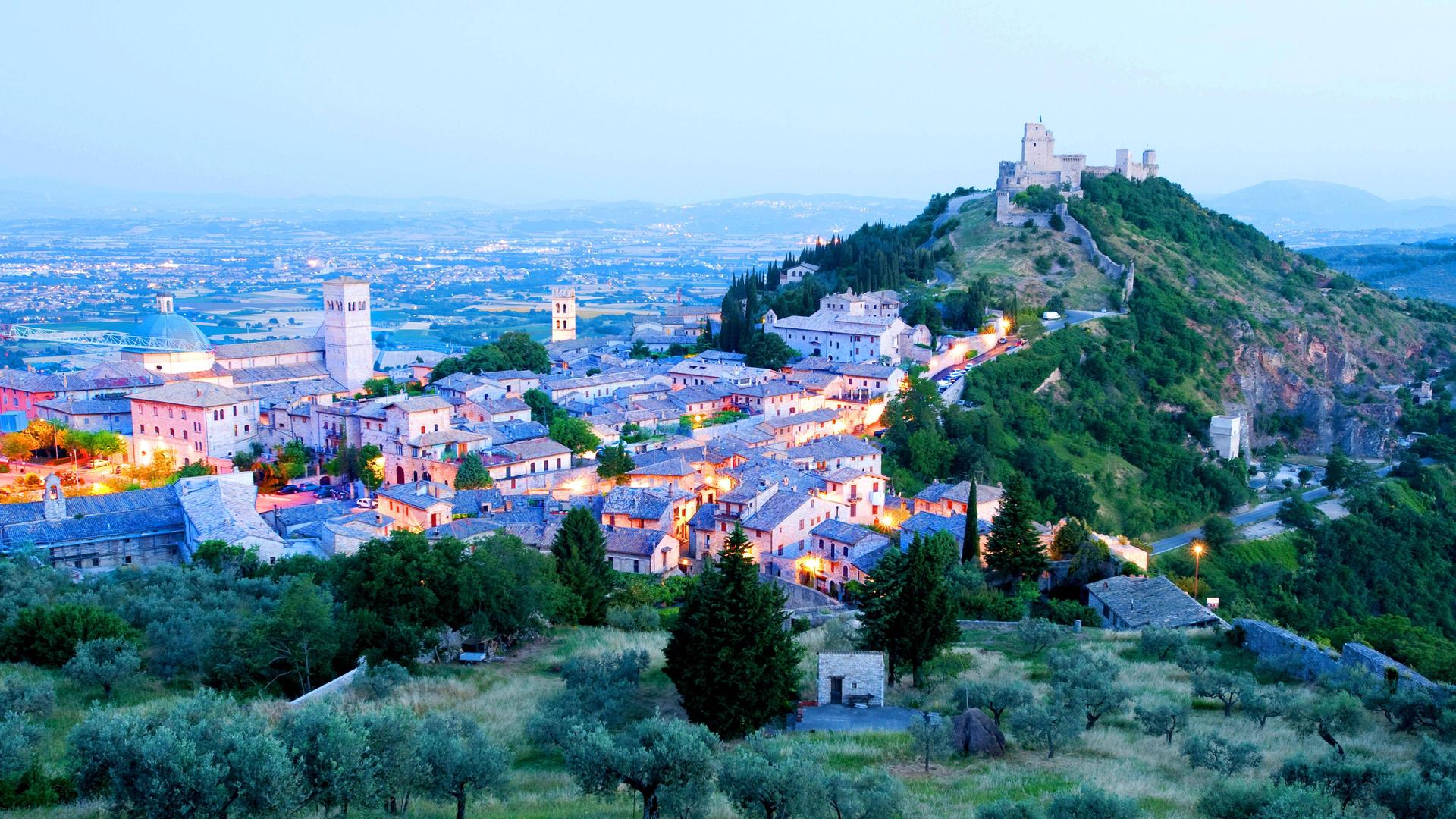 Panoramablick über die italienische Stadt Assisi in Region Umbrien. Das Landschaftsbild wird von einer mittelalterlichen Burg auf einem Hügel dominiert. In der Stadt leuchten Lichter.