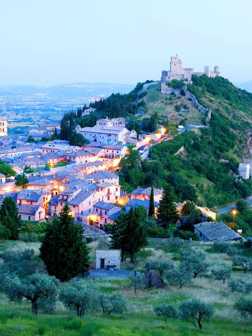 Panoramablick über die italienische Stadt Assisi in Region Umbrien. Das Landschaftsbild wird von einer mittelalterlichen Burg auf einem Hügel dominiert. In der Stadt leuchten Lichter. Panoramablick über die italienische Stadt Assisi in Region Umbrien. Das Landschaftsbild wird von einer mittelalterlichen Burg auf einem Hügel dominiert. In der Stadt leuchten Lichter.