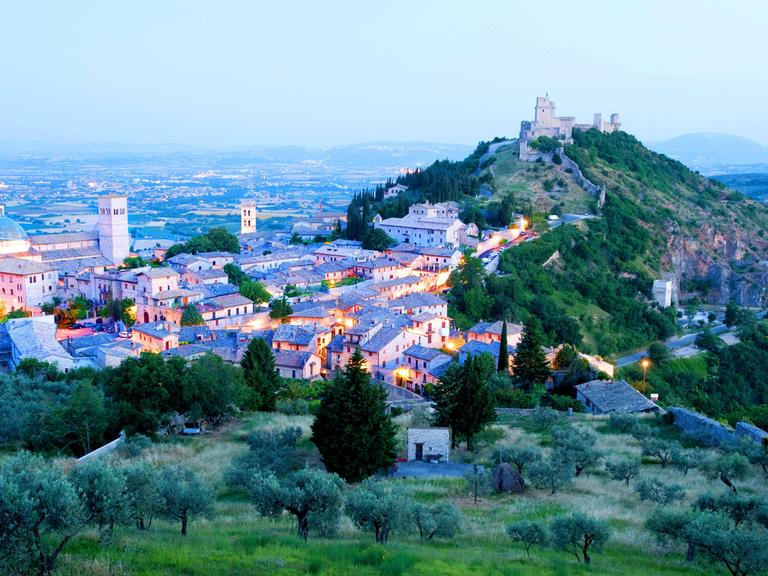 Panoramablick über die italienische Stadt Assisi in Region Umbrien. Das Landschaftsbild wird von einer mittelalterlichen Burg auf einem Hügel dominiert. In der Stadt leuchten Lichter. Panoramablick über die italienische Stadt Assisi in Region Umbrien. Das Landschaftsbild wird von einer mittelalterlichen Burg auf einem Hügel dominiert. In der Stadt leuchten Lichter.
