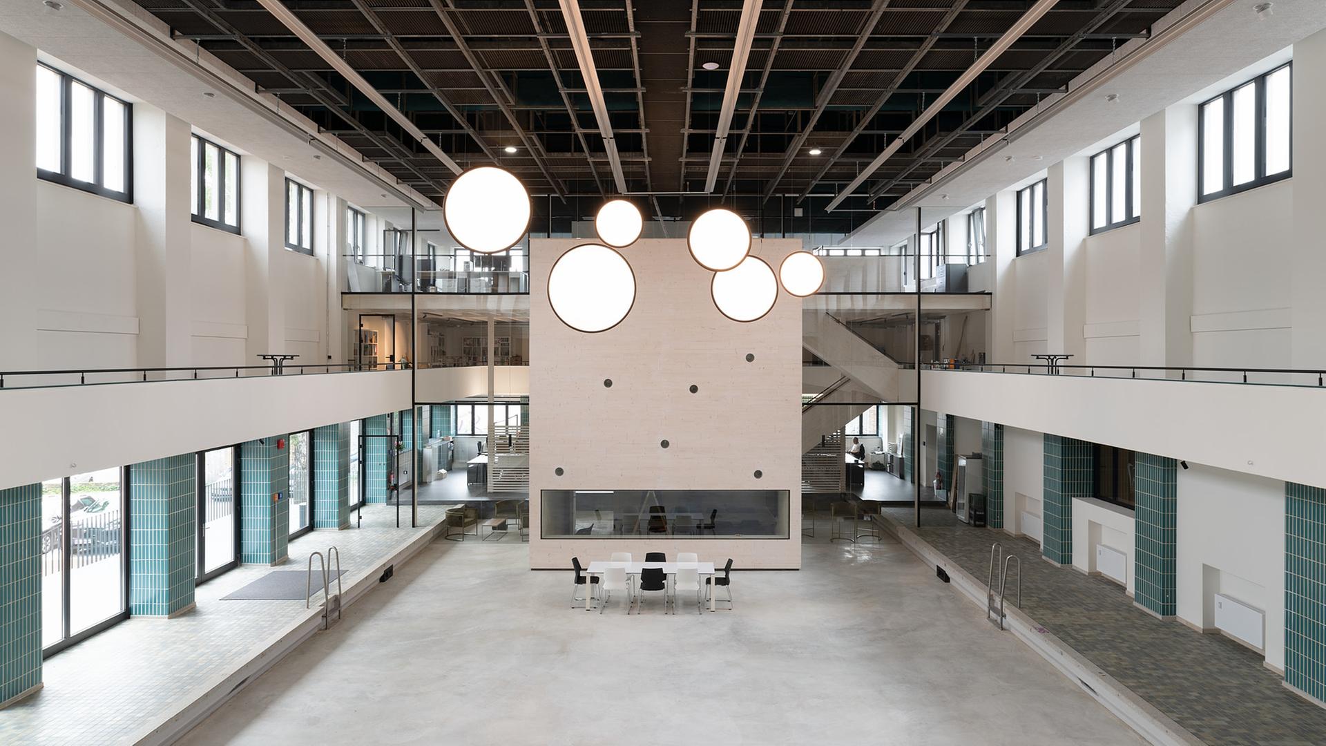 Blick in eine ehemalige Schwimmhalle: im leeren Becken ein quaderförmiger Pavillon, von der Decke hängt eine Installation mit kreisförmigen, leuchtend weißen Scheiben.