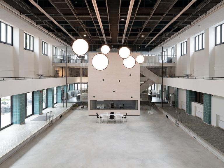 Blick in eine ehemalige Schwimmhalle: im leeren Becken ein quaderförmiger Pavillon, von der Decke hängt eine Installation mit kreisförmigen, leuchtend weißen Scheiben.