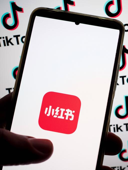 Auf einem Smartphone ist das Logo der Rednote-App zu sehen. Im Hintergrund prangen TikTok-Logos auf einem Bildschirm.