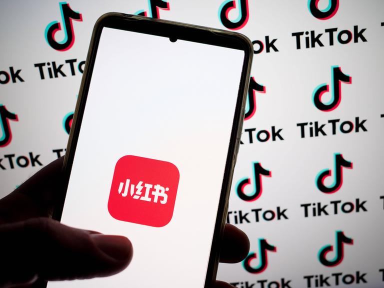 Auf einem Smartphone ist das Logo der Rednote-App zu sehen. Im Hintergrund prangen TikTok-Logos auf einem Bildschirm.