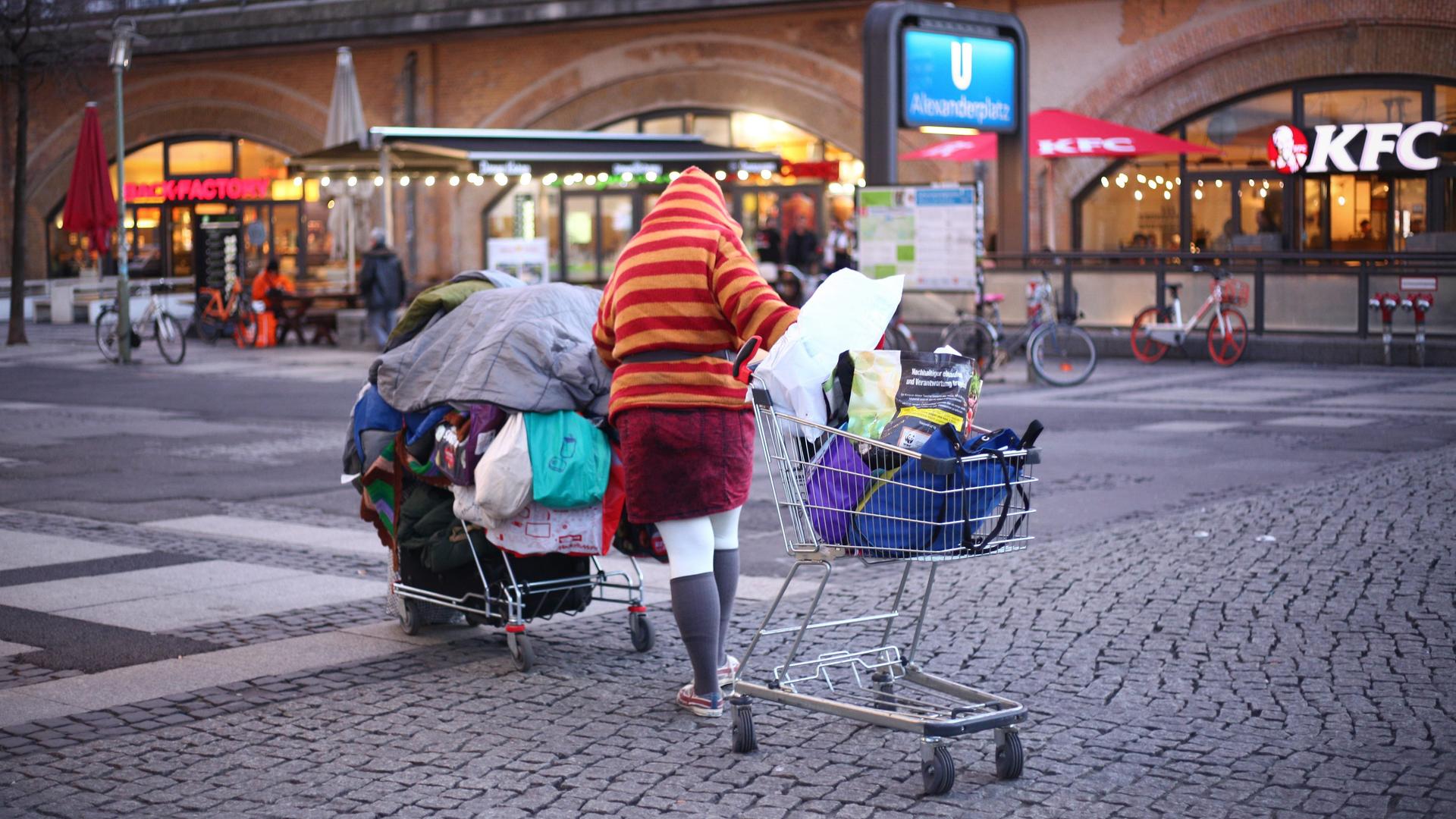 Eine obdachlose Person schiebt im Bezirk Mitte auf dem Alexanderplatz bei einbrechender Dämmerung ihre Habe in zwei Einkaufswagen über den Platz.