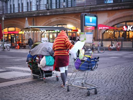 Eine obdachlose Person schiebt im Bezirk Mitte auf dem Alexanderplatz bei einbrechender Dämmerung ihre Habe in zwei Einkaufswagen über den Platz.