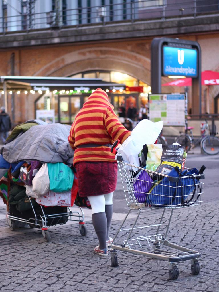 Eine obdachlose Person schiebt im Bezirk Mitte auf dem Alexanderplatz bei einbrechender Dämmerung ihre Habe in zwei Einkaufswagen über den Platz.