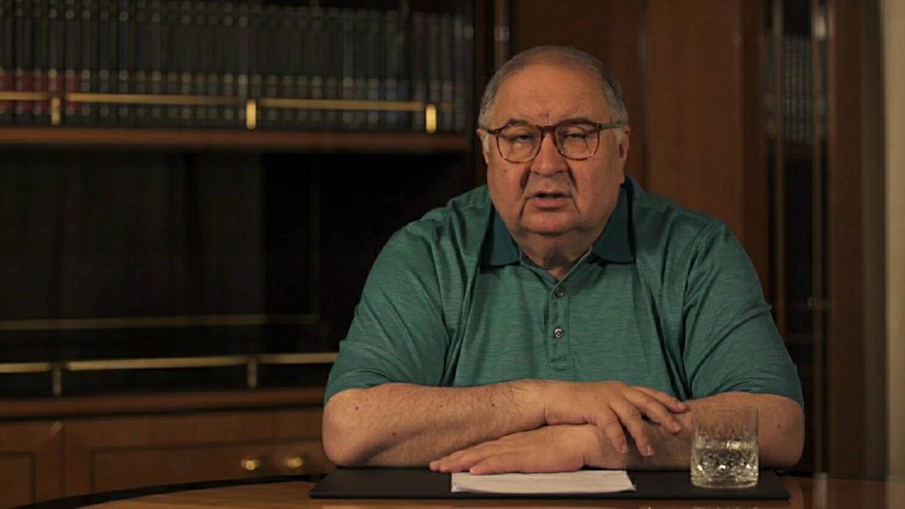 Der russische Milliardär Alischer Usmanow sitzt an einem Schreibtisch und schaut in die Kamera, seine Hände sind aufeinandergelegt. 