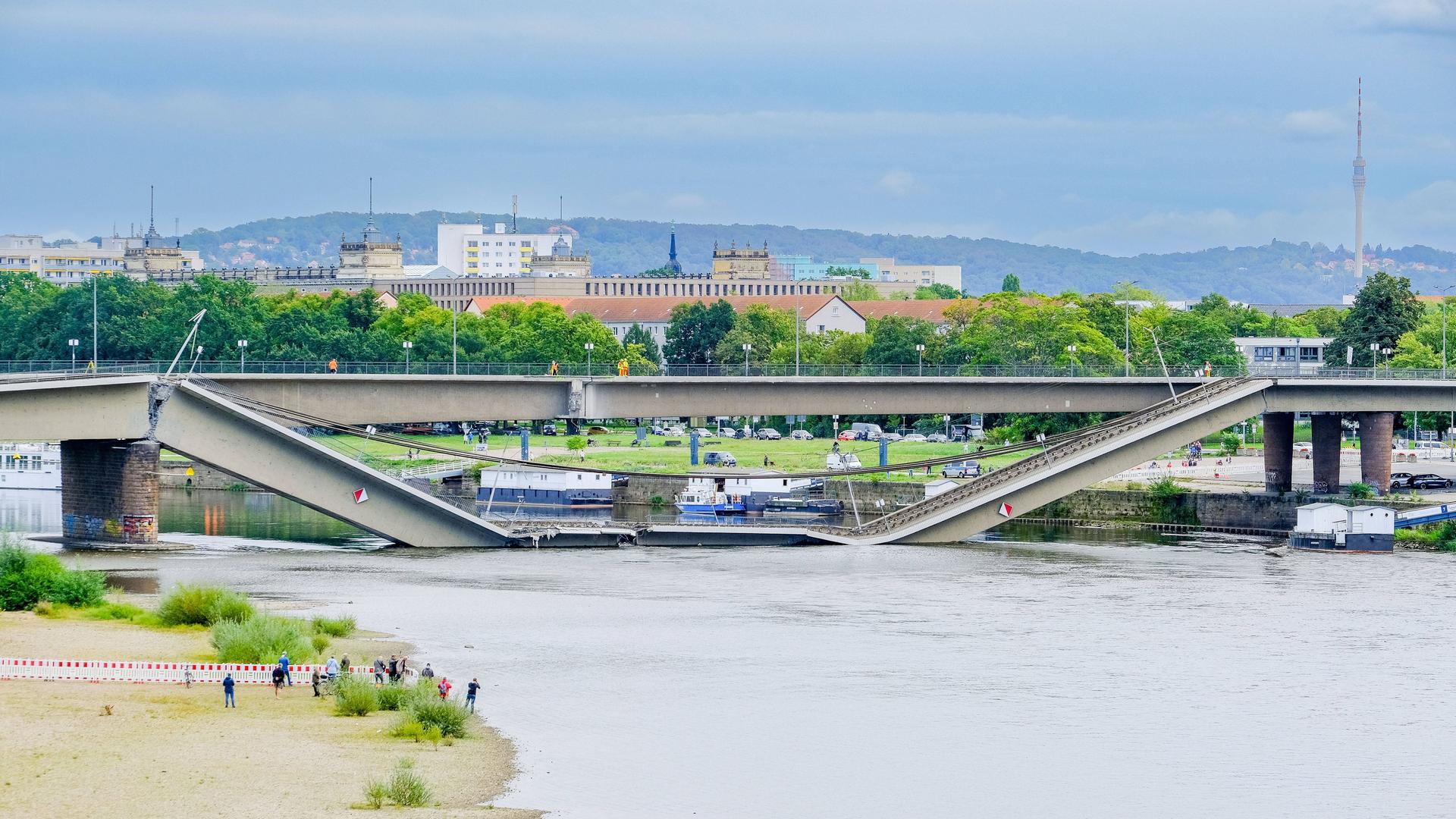 Blick auf die Elbe in Dresden mit dem eingestürzte Brückenzug der Carolabrücke. Vorne sind am Elbstrand mehrere Menschen zu sehen.