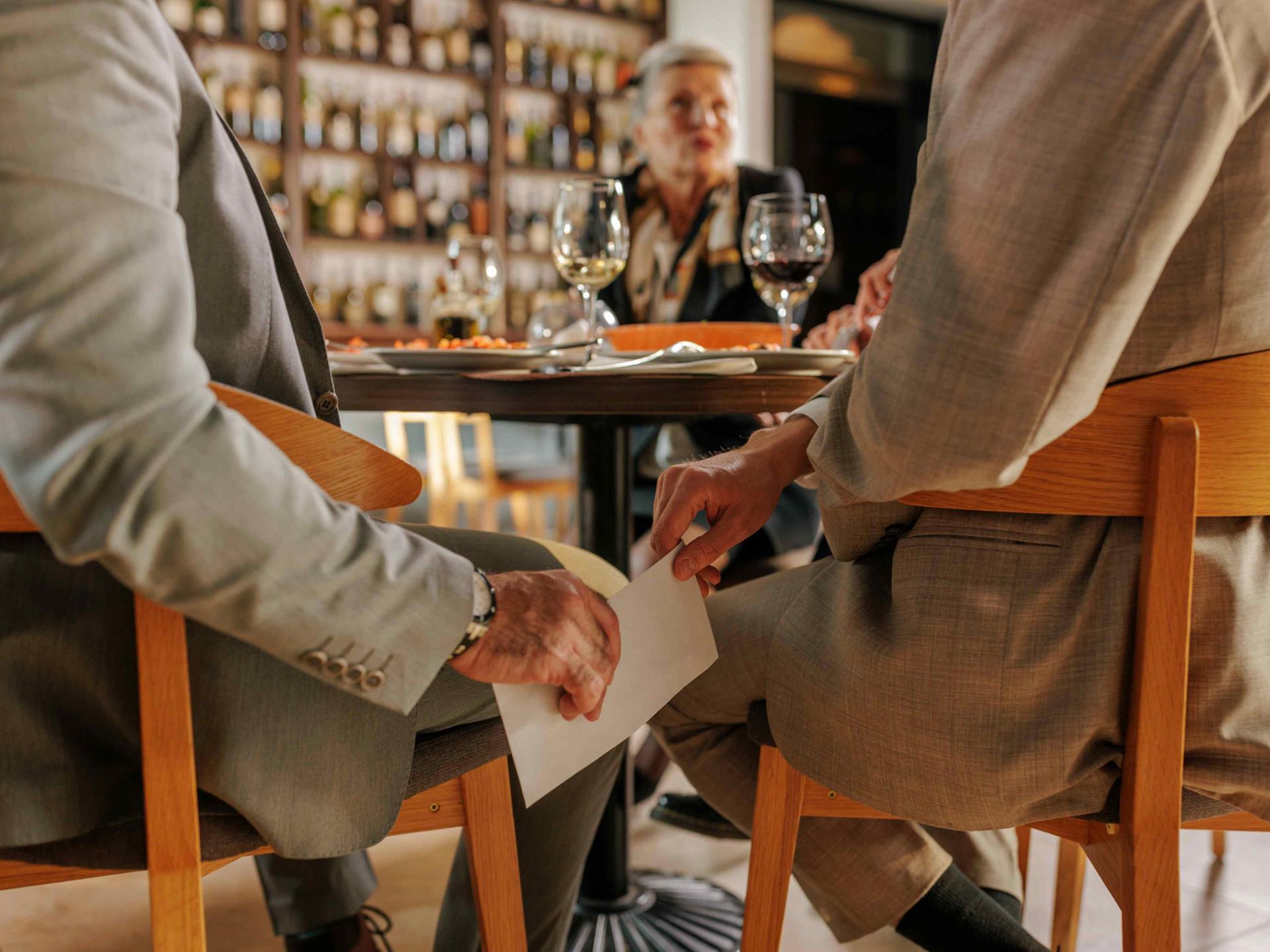 In einem Restaurant reicht ein Mann dem anderen unter dem Tisch einen weißen Umschlag.