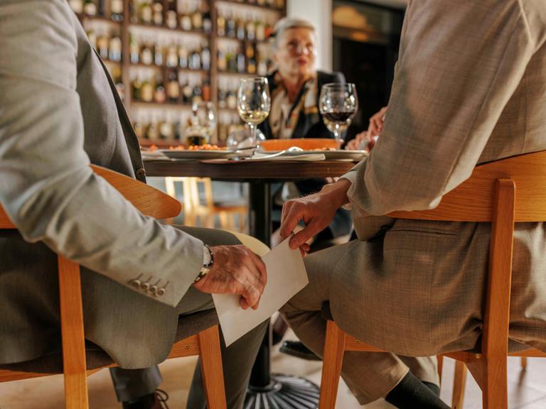 In einem Restaurant reicht ein Mann dem anderen unter dem Tisch einen weißen Umschlag. In einem Restaurant reicht ein Mann dem anderen unter dem Tisch einen weißen Umschlag.