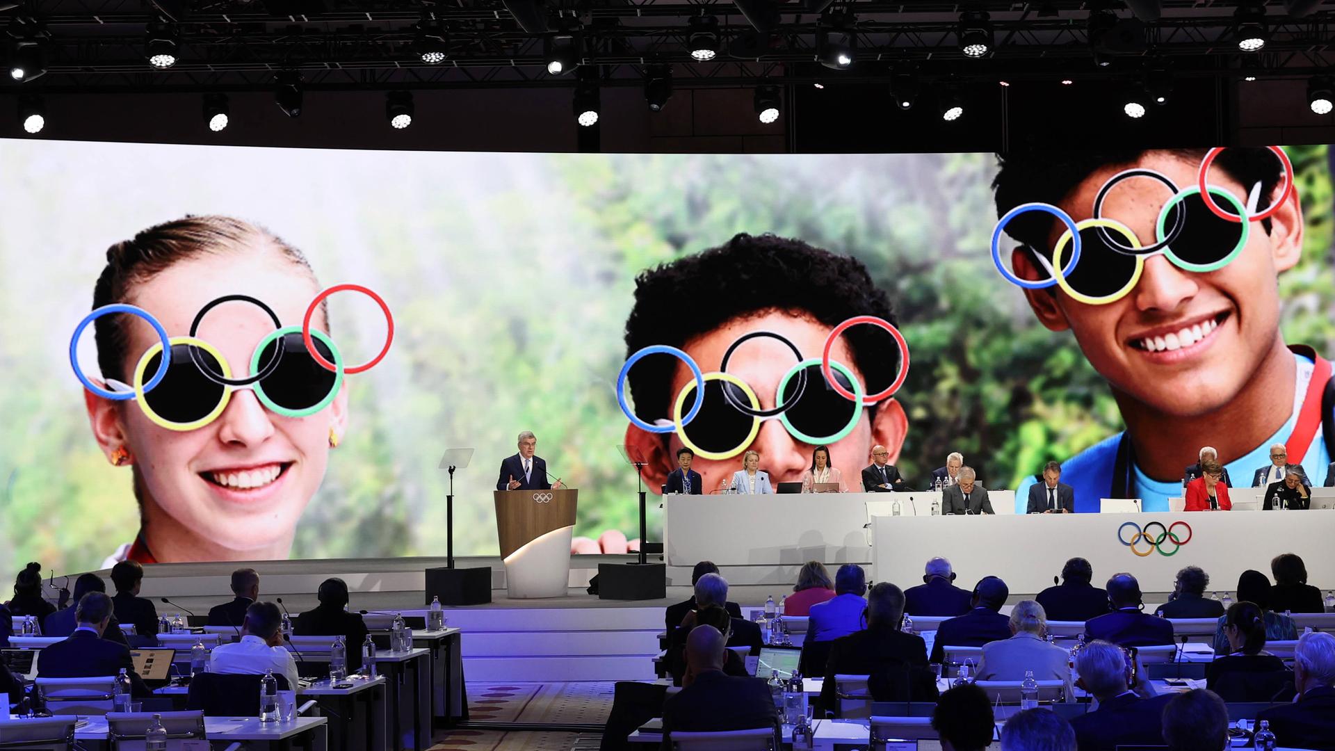 Thomas Bach spricht bei der IOC-Session in Griechenland vor Teilnehmern, im Hintergrund sind junge Menschen mit Sonnenbrillen mit olympischen Ringen abgebildet. Thomas Bach spricht bei der IOC-Session in Griechenland vor Teilnehmern, im Hintergrund sind junge Menschen mit Sonnenbrillen mit olympischen Ringen abgebildet.