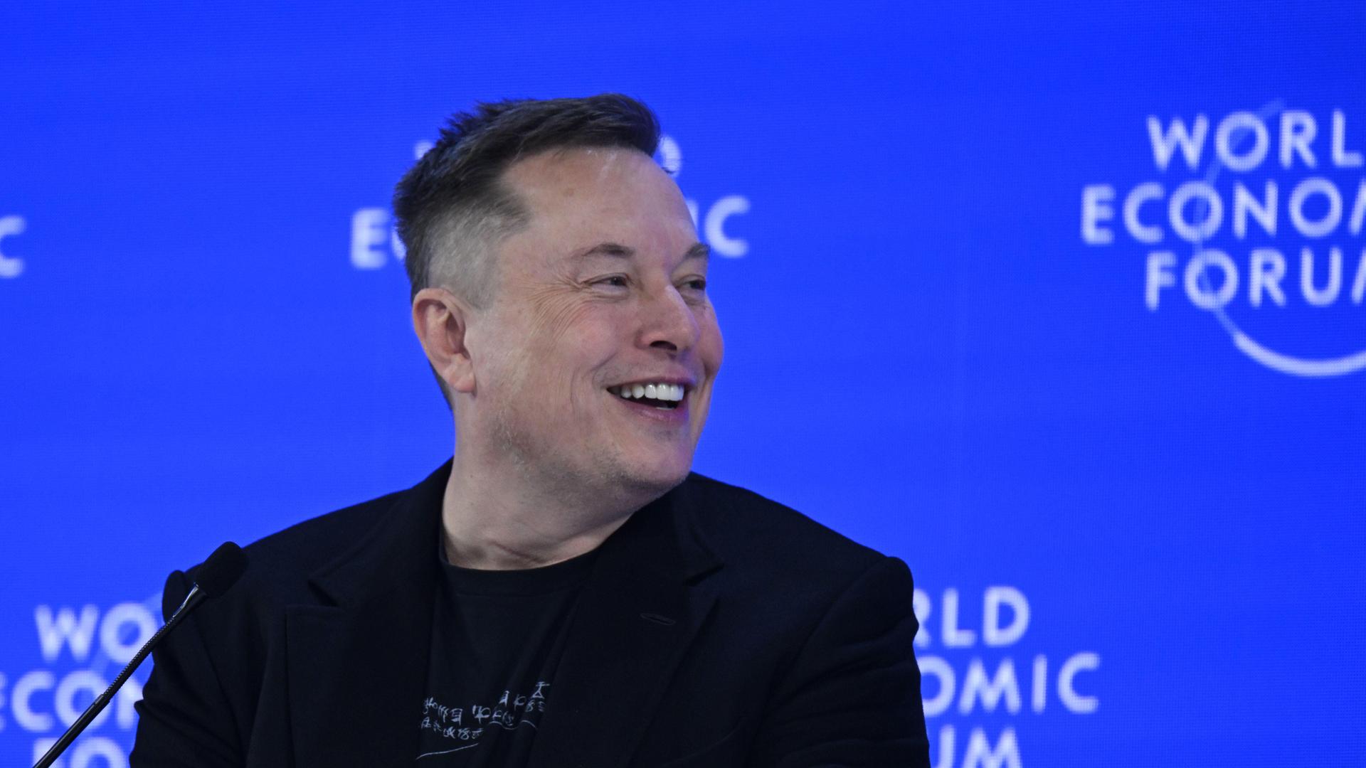 Elon Musk sitzt vor einer Logo-Tapete des Weltwirtschaftsforums und lächelt aus dem Bild heraus.