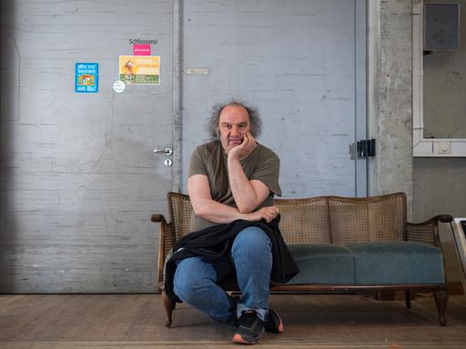 Matthias Lilienthal, Ex-Intendant der Münchner Kammerspiele, sitzt vor einer kargen Wand auf einem Sofa, das Kinn auf eine Hand gestützt.