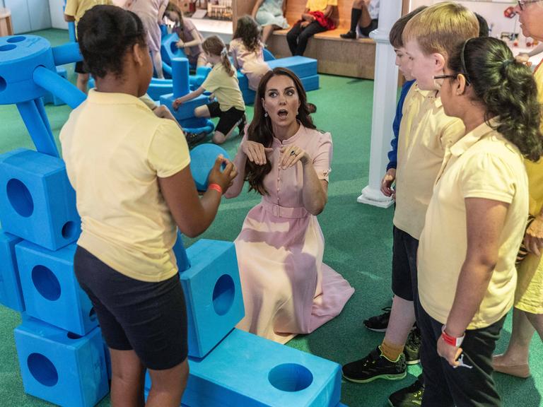 Prinzessin Kate kniet in einer Gruppe von Kindern, daneben große blaue Spielelemente für Kinder.