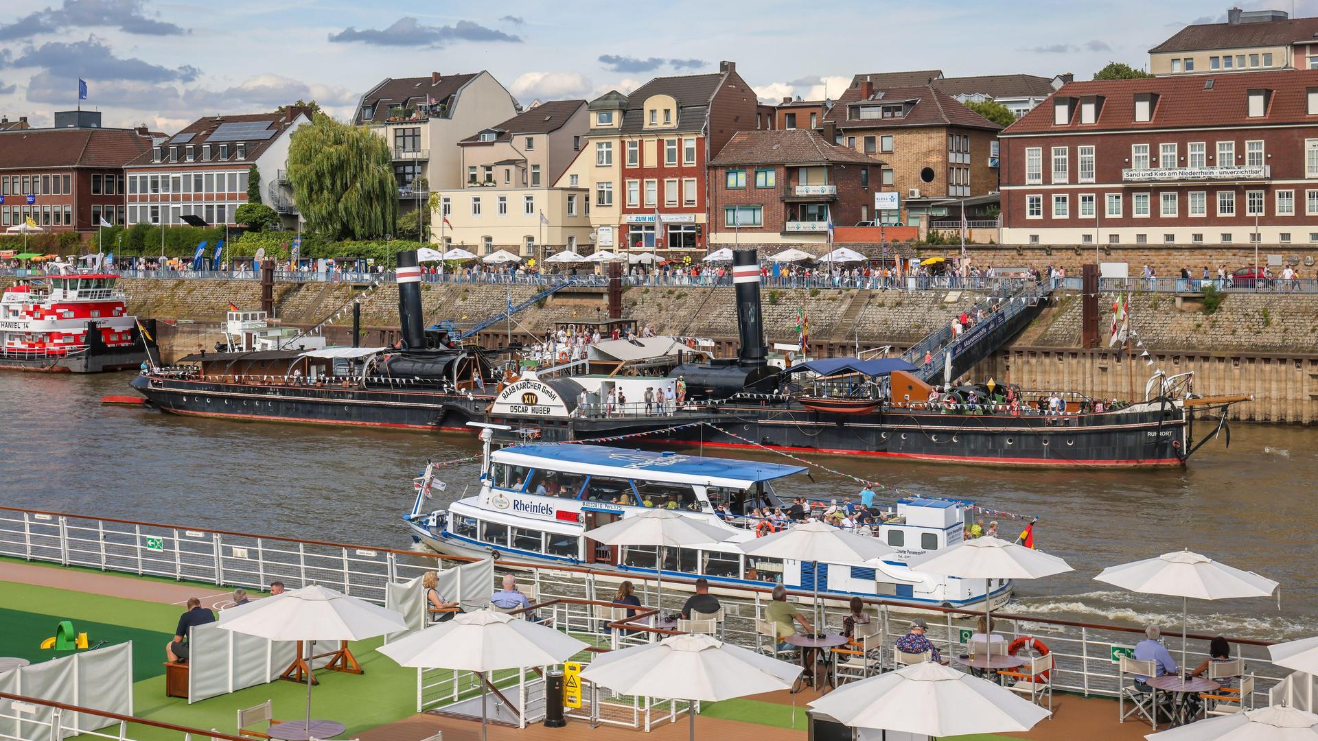 Bei einem Hafenfest liegen Flusskreuzfahrt- und Museumsschiffe im Hafen. Im Bildhintergrund sind Wohnhäuser zu sehen.
