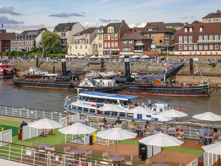 Bei einem Hafenfest liegen Flusskreuzfahrt- und Museumsschiffe im Hafen. Im Bildhintergrund sind Wohnhäuser zu sehen.