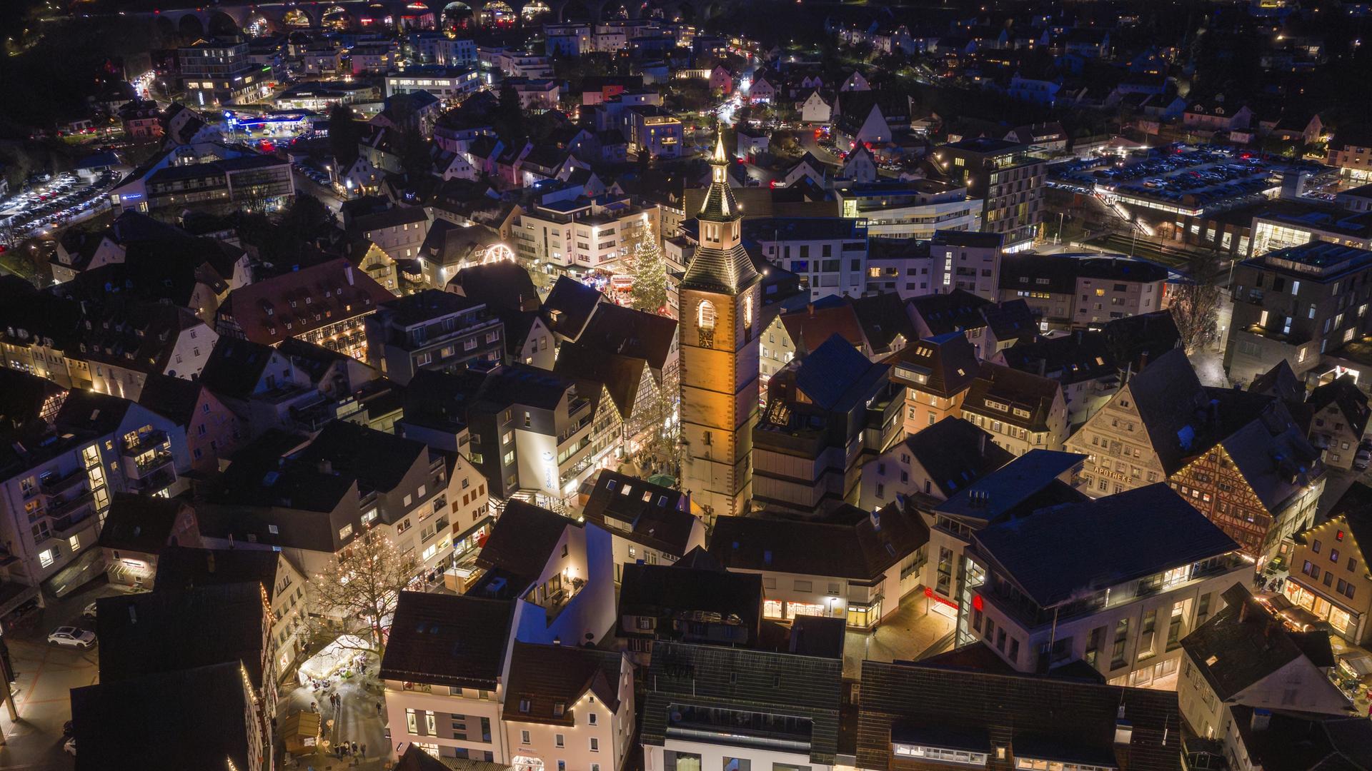 Luftaufnahme einer beleuchteten Stadt bei Nacht, dominiert von einer großen Kirche, umgeben von festlich beleuchteten Häusern.