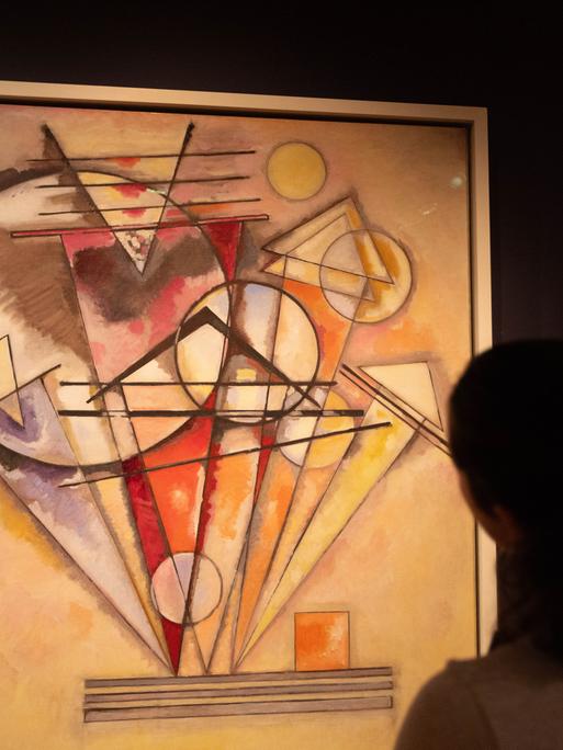 Zu sehen ist ein abstraktes Bild, das Wassily Kandinsky gemalt hat. Wesentliche Elemente darin sind Kreise und Dreiecke. Der Hintergrund des Kunstwerks ist in Beigetönen gehalten. Das Hauptmotiv erscheint in den Farben Rot, Orange, Gelb und Violett.