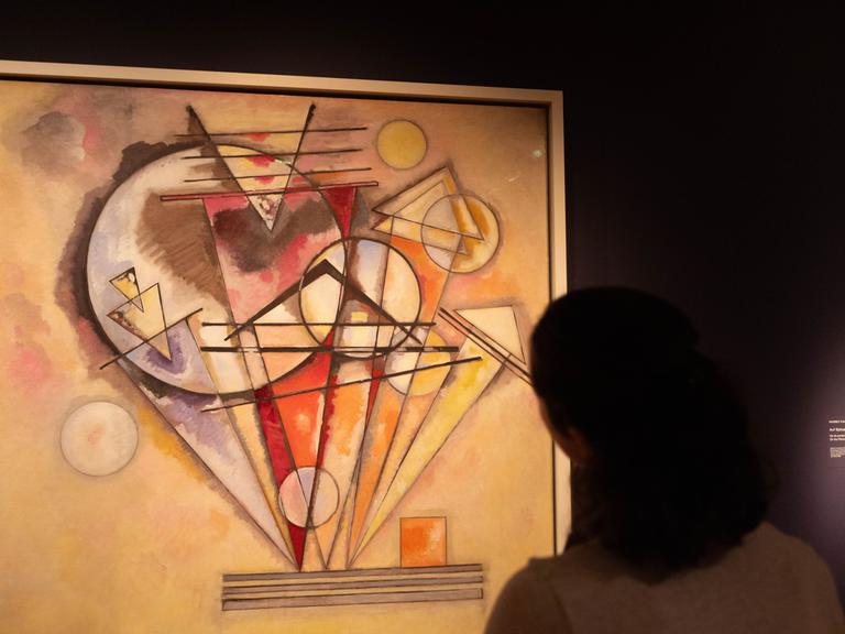 Zu sehen ist ein abstraktes Bild, das Wassily Kandinsky gemalt hat. Wesentliche Elemente darin sind Kreise und Dreiecke. Der Hintergrund des Kunstwerks ist in Beigetönen gehalten. Das Hauptmotiv erscheint in den Farben Rot, Orange, Gelb und Violett.