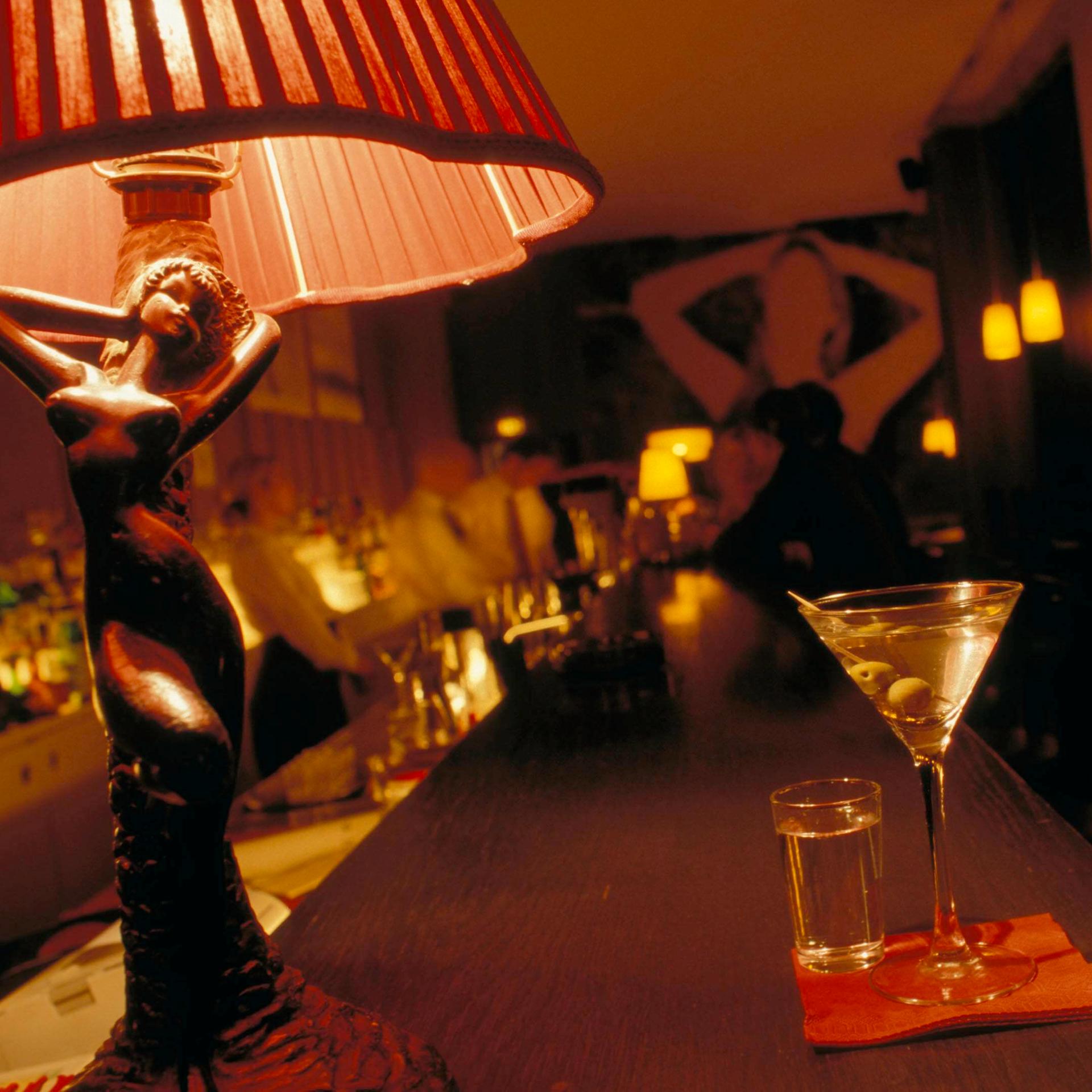 Auf einem Tresen in einer Bar steht ein Martiniglas. Daneben eine Tischlampe, deren hölzerner Sockel die Form einer Frau hat.