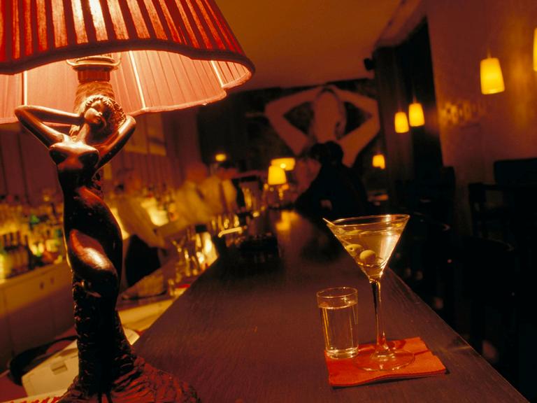 Auf einem Tresen in einer Bar steht ein Martiniglas. Daneben eine Tischlampe, deren hölzerner Sockel die Form einer Frau hat.
