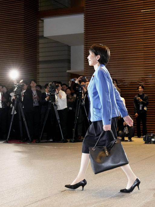 Sanae Takaichi läuft im Businessoutfit und mit Aktentasche an einer langen Reihe Fotografen vorbei.