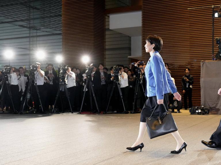 Sanae Takaichi läuft im Businessoutfit und mit Aktentasche an einer langen Reihe Fotografen vorbei.