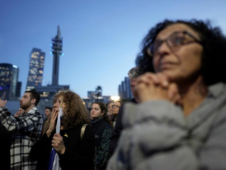 Menschen stehen vor Hochhäusern beieinander. Eine Frau im Vordergrund hat die Hände wie für ein Gebet gefaltet. Es sind Angehörige von Geiseln der Hamas.