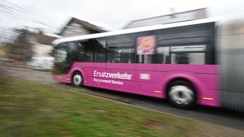 Ein lilafarbener Bus mit der Aufschrift „Ersatzverkehr – Replacement Service“ fährt auf einer Straße. Im Hintergrund sind Wohnhäuser zu sehen.