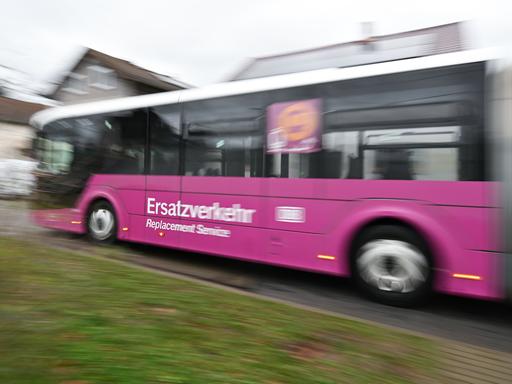 Ein lilafarbener Bus mit der Aufschrift „Ersatzverkehr – Replacement Service“ fährt auf einer Straße. Im Hintergrund sind Wohnhäuser zu sehen.
