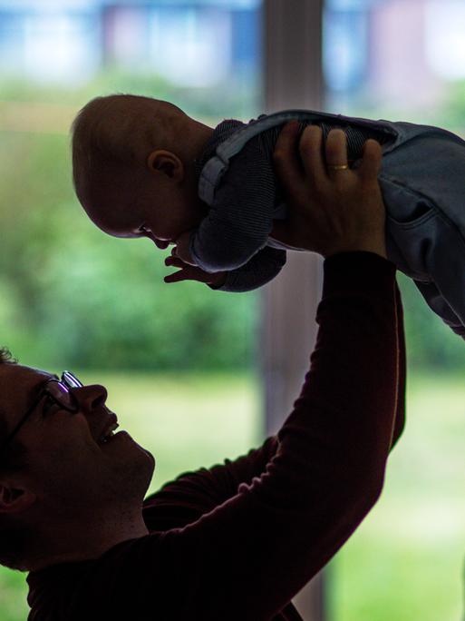 Ein junger Vater spielt mit seinem sechs Monate alten Sohn im Wohnzimmer, hält ihn in die Luft, der Säugling lacht