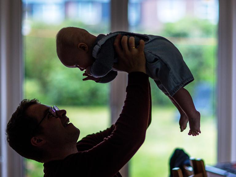 Ein junger Vater spielt mit seinem sechs Monate alten Sohn im Wohnzimmer, hält ihn in die Luft, der Säugling lacht