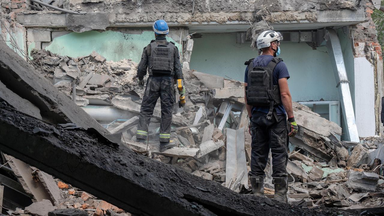 Ein zerstörtes Gebäude in der Ukraine. Auf den Trümmern stehen zwei Männer mit Schutzhelmen. 