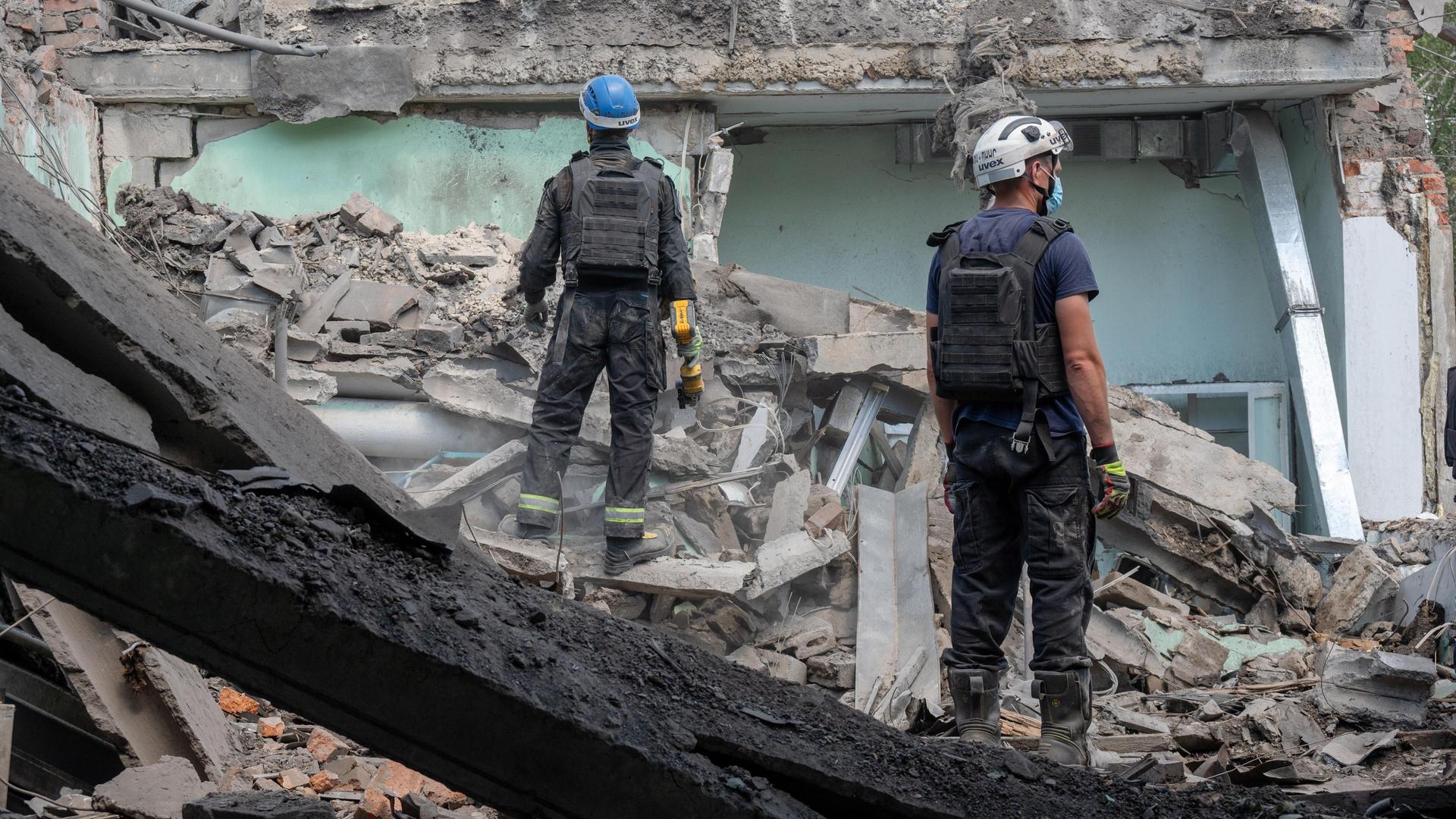 Ein zerstörtes Gebäude in der Ukraine. Auf den Trümmern stehen zwei Männer mit Schutzhelmen. 
