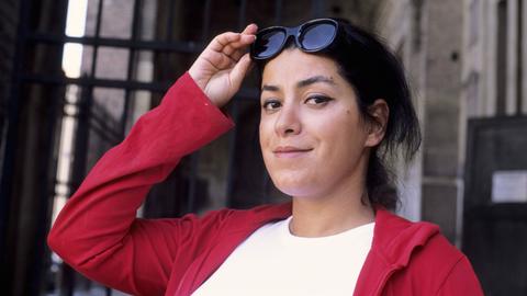 Die Künstlerin Marjane Satrapi im Porträt. Sie schaut verschmitzt in die Kamera, hält ihre Sonnenbrille fest und trägt eine rote Jacke