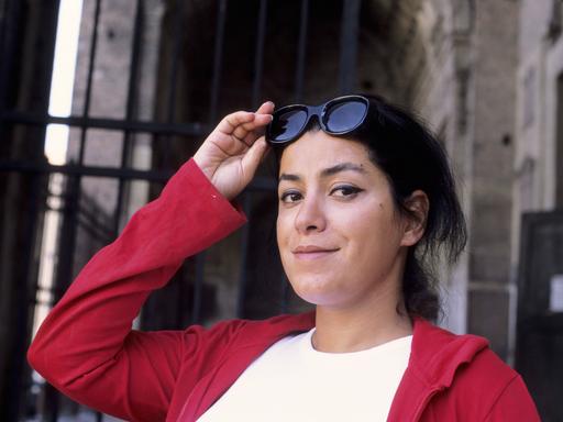 Die Künstlerin Marjane Satrapi im Porträt. Sie schaut verschmitzt in die Kamera, hält ihre Sonnenbrille fest und trägt eine rote Jacke