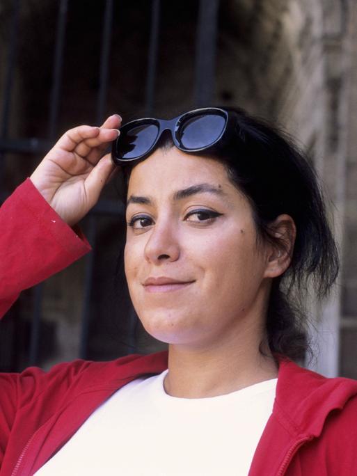 Die Künstlerin Marjane Satrapi im Porträt. Sie schaut verschmitzt in die Kamera, hält ihre Sonnenbrille fest und trägt eine rote Jacke